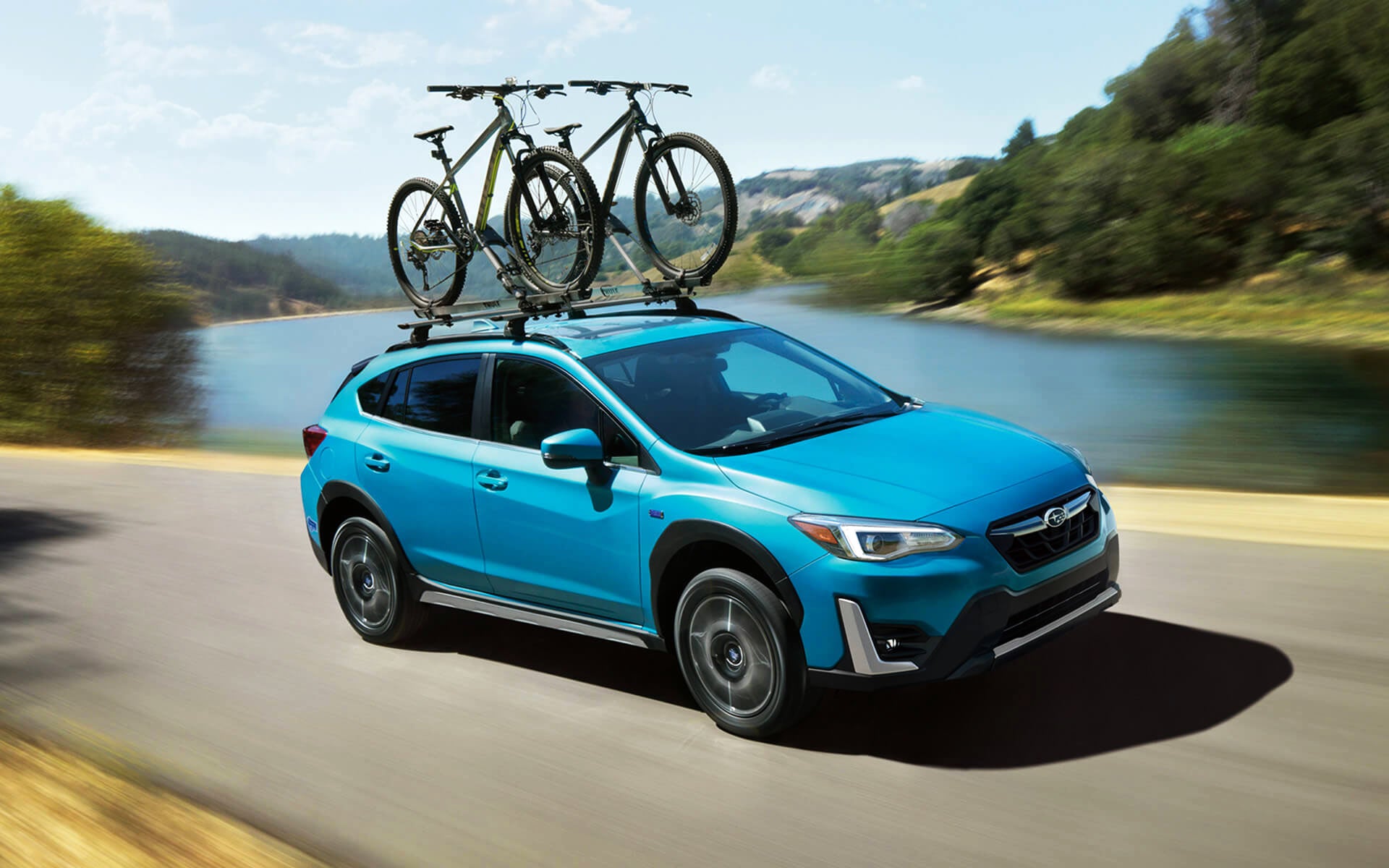 A blue Crosstrek Hybrid with two bicycles on its roof rack driving beside a river | Subaru World of Newton in Newton NJ